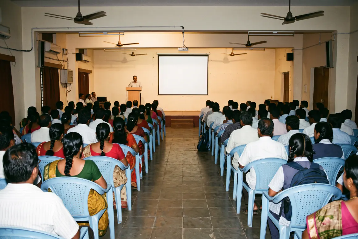 A hall of seated attendees facing a speaker at a lectern beside a projection screen.